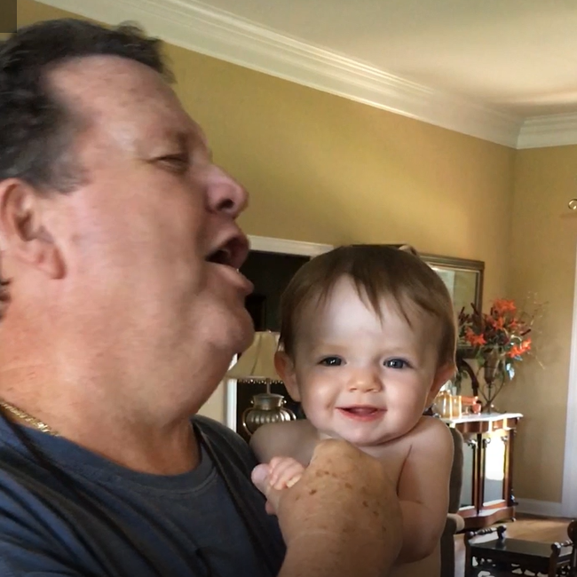 Keith Lowhorne holds his grandchild and seems to be singing and dancing. The baby is looking right at the camera and smiling.