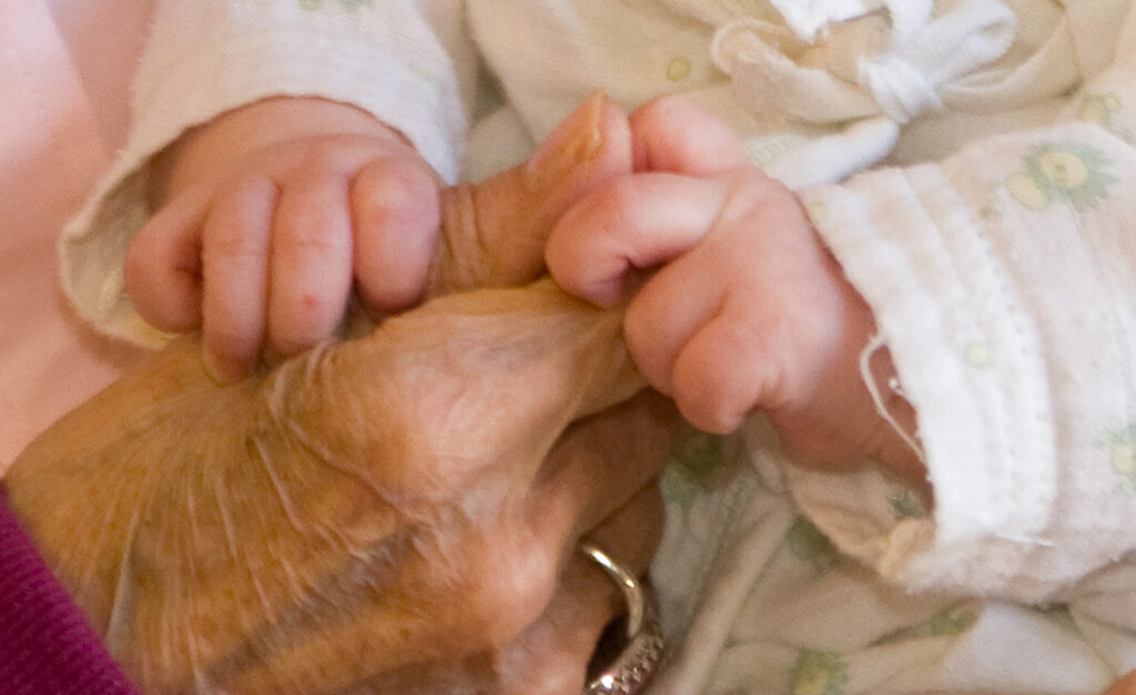 A baby's hands grasp the hand of an older person.