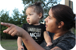 A Native toddler and adult together outside. The toddler's gaze follows the adult's pointed finger, while the adult looks at the child.