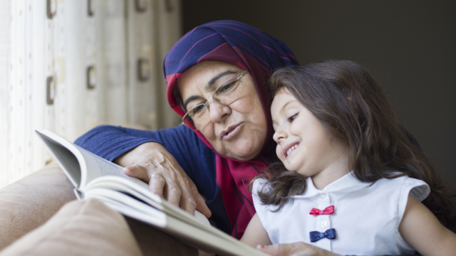 A grandmother in a headscarf reads with her young granddaughter, who is smiling