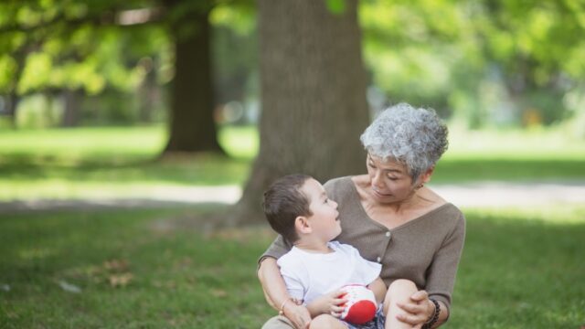 An older woman sits on the ground in a park and holds her young grandson in her lap. They two are looking at each other intently and happily. Both are of Asian descent.