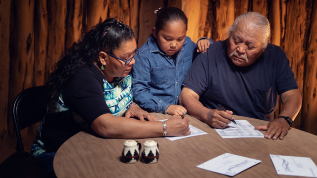 A Native child stands between two older Native adults as they sit at a table and fill out paperwork