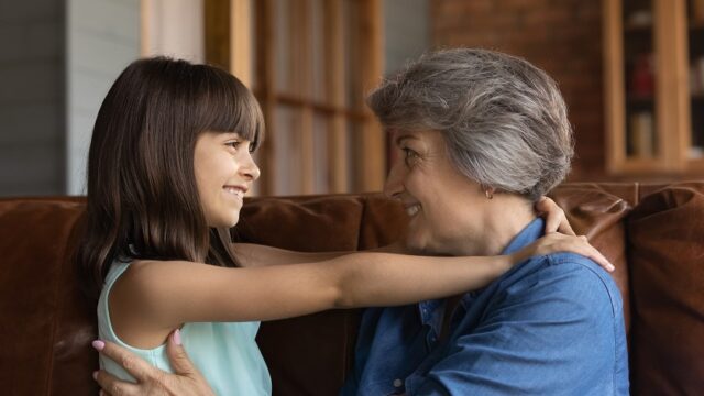 A Latina grandmother and her elementary-aged granddaughter smile at each other with their arms around each other