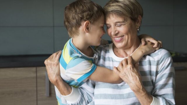 A white grandmother smiles and closes her eyes as she holds onto her grandson, who is smiling and embracing her from the side