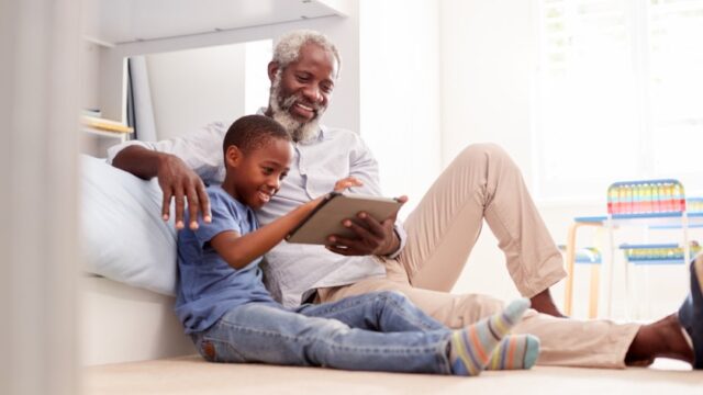 A Black grandfather and his grandson sit on the floor together and smile as they look at a tablet together