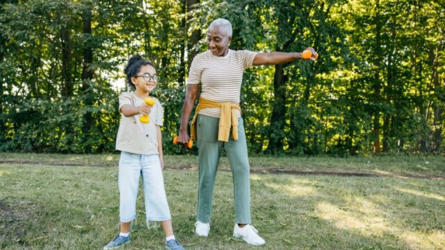 An African American grandmother smiles at her smiling granddaughter as they lift dumbbells together in a park