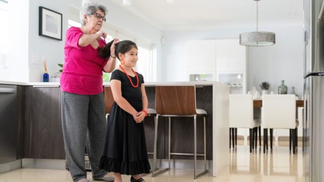 A girl in a fancy dress smiles as her grandmother does her hair. The family is Cuban-American.