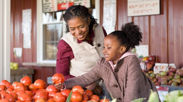 A Black grandmother and granddaughter smile as they pick out fruits at a farm stand