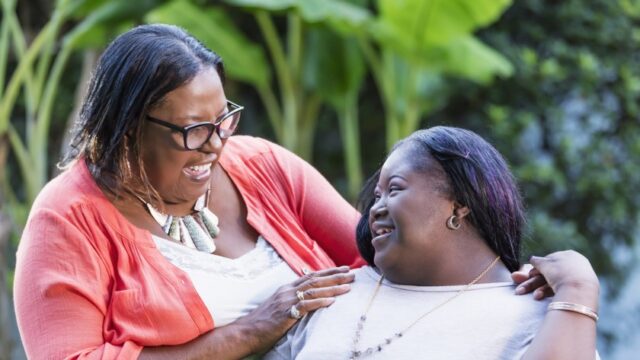 An African-American woman in her 50s standing with her teenage granddaughter, 16 years old, who has Down Syndrome. They are outdoors, in the back yard, smiling at each other.