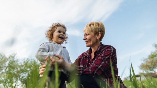 A white grandmother and her young grandchild stand in a field together. The grandmother is sitting or kneeling and looking at and holding onto her grandchild and the child is smiling.