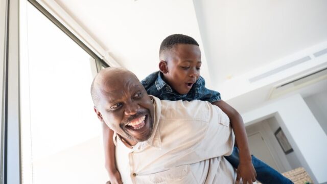 A Black grandfather smiles widely as he plays with his young grandson, holding the joyful child on his back