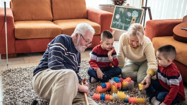 A white grandmother and grandfather sit on the floor with their twin grandsons, playing with large, Lego-type blocks